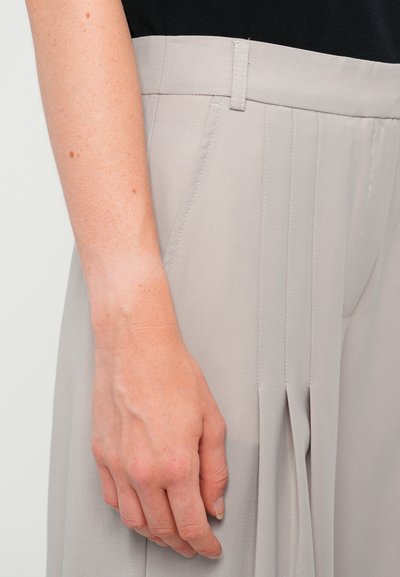 Lichtgrijze geplooide broek met voorzakken, een gladde textuur en subtiele stikseldetails, gedragen met een zwarte top, waarin een hand te zien is.
