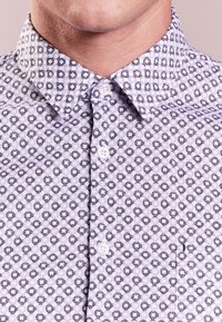 Světle fialová košile s geometrickým černobílým vzorem s hexagonálními tvary. Design s límečkem a bílými knoflíky, hladká struktura látky.