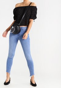 Schwarzes Top mit Punkten und schulterfrei, blaue Skinny-Jeans mit ausgefranstem Saum, schwarze Handtasche mit runden Beschlägen und schwarze spitze Absätze.