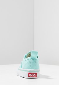 Vaaleansininen kangaskenkä valkoisella kumipohjalla, jossa on pieni kantatappi ja punainen Vans-logo kantapäässä. Yksinkertainen, matalahko muotoilu.