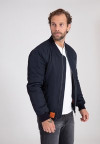 Giacca bombardiere della marina con polsini e colletto a costine, chiusura con zip, tasche laterali e un'etichetta arancione sull'orlo. Indossata sopra una camicia bianca.