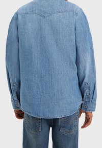 Osoba oblečená v světle modré džínsové košili a středně modrých džínách, zobrazená zezadu na jednobarevném pozadí.