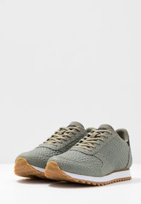 Vihreät teksturoidut juoksukengät, joissa on matomaista kuviota, vaaleanbeige narut, valkoinen välikiila ja ruskea kumipohja.