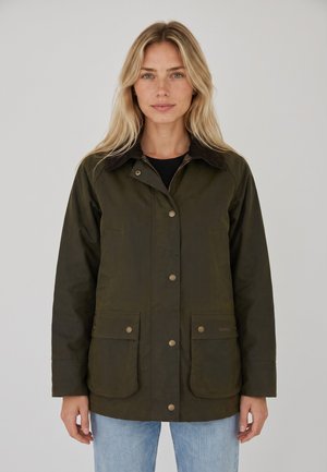 Femme blonde portant une veste vert olive avec des poches avant boutonnées et un col foncé, debout devant un fond clair uni.