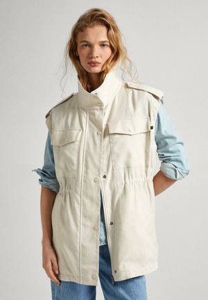 Chaqueta sin mangas beige con un alto cuello, dos bolsillos en el pecho y detalle de cintura elástica. Lleva puesta una camisa de color azul claro. Diseño casual.