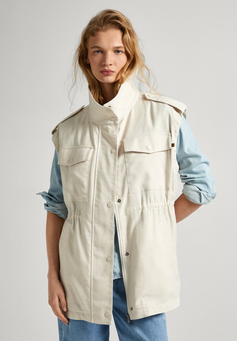 Chaqueta sin mangas beige con un alto cuello, dos bolsillos en el pecho y detalle de cintura elástica. Lleva puesta una camisa de color azul claro. Diseño casual.