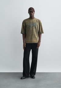 Palaidi alyvuogių žalia marškinėliai su spauda "ALAMO GODS", derinami su juodomis plačiomis kelnėmis ir juodais batais, užsidėti su ruda kepure.