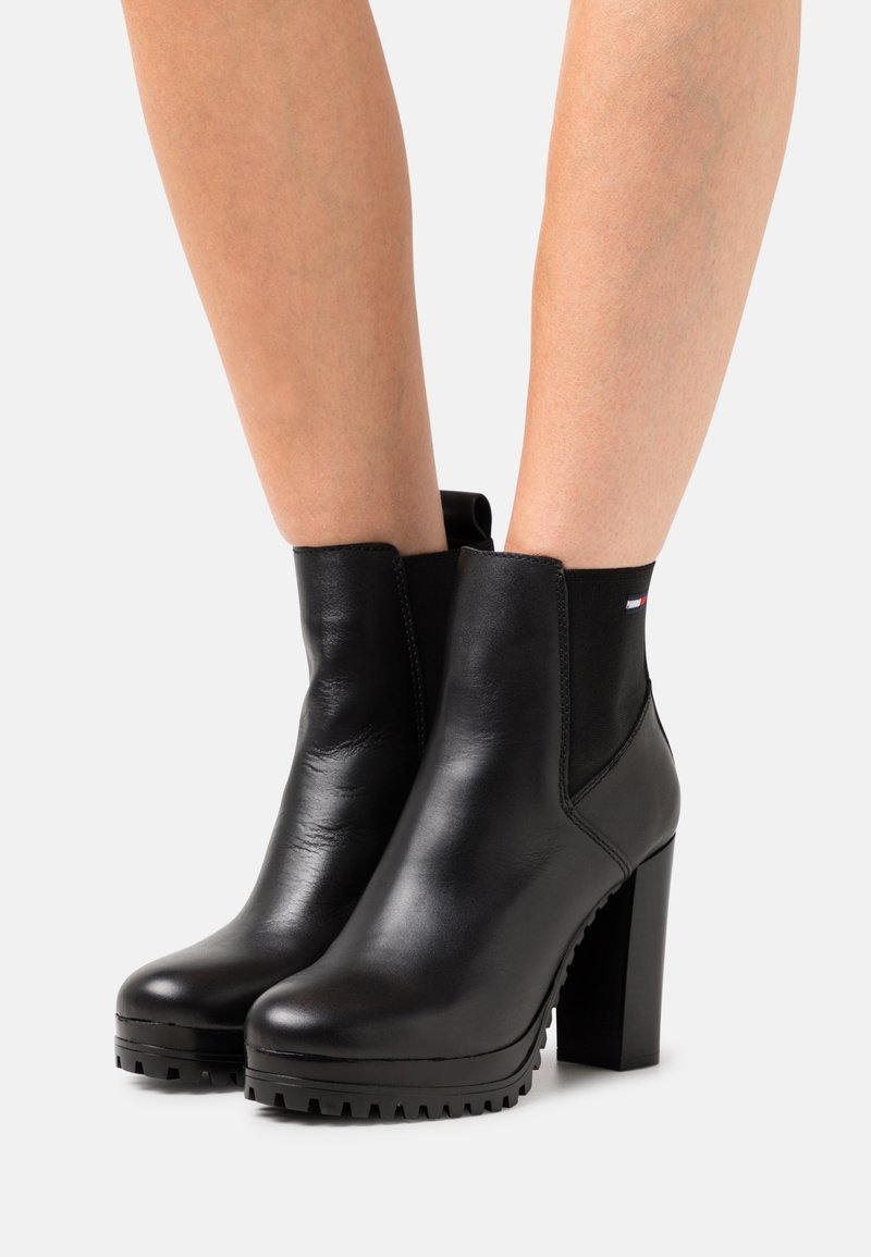 Bottines en cuir noir avec talons hauts épais, panneaux latéraux élastiques et semelles à crampons portées sur jambes nues sur fond blanc.