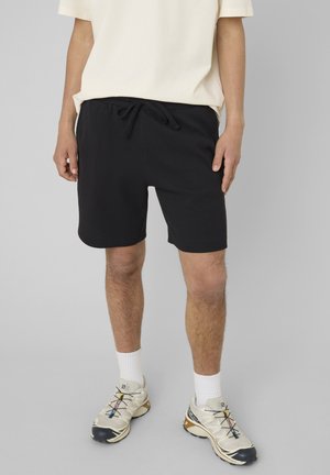 Personne portant un short noir à cordon, des chaussettes blanches, des chaussures de sport beige et noires, et un t-shirt crème à manches courtes, debout sur un fond gris.
