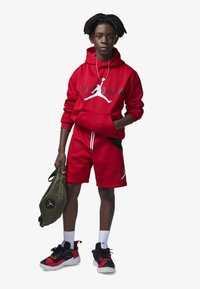 Sudadera roja con el logo de "Jordan" y bolsillo tipo canguro, combinada con pantalones cortos rojos. Zapatillas deportivas negras y rojas, bolsa verde con detalles del logo.