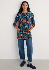 Ženska v barvni tuniki z dolgimi rokavi in žepi, z modrimi kavbojkami s poševnimi rokavi in mornarskimi supergami, ki stoji ob svetli enobarvni steni.