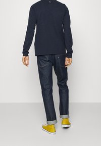 Osoba oblečená v tmavě modré košili s dlouhým rukávem, tmavých vyhrnutých džínách a žlutých teniskách Converse na vysoké platformě stojí proti jednoduchému světlému pozadí.