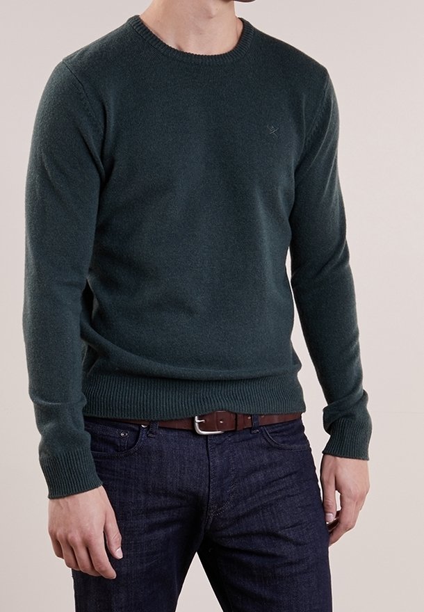 Homme portant un pull ras du cou vert foncé, un jean bleu foncé, et une ceinture en cuir marron, debout devant un fond uni.