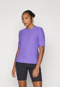 Chemise violette à manches courtes avec un col rond, texture lisse et design ajusté. Associée à un short noir, montrant des bras à la teinte de peau.