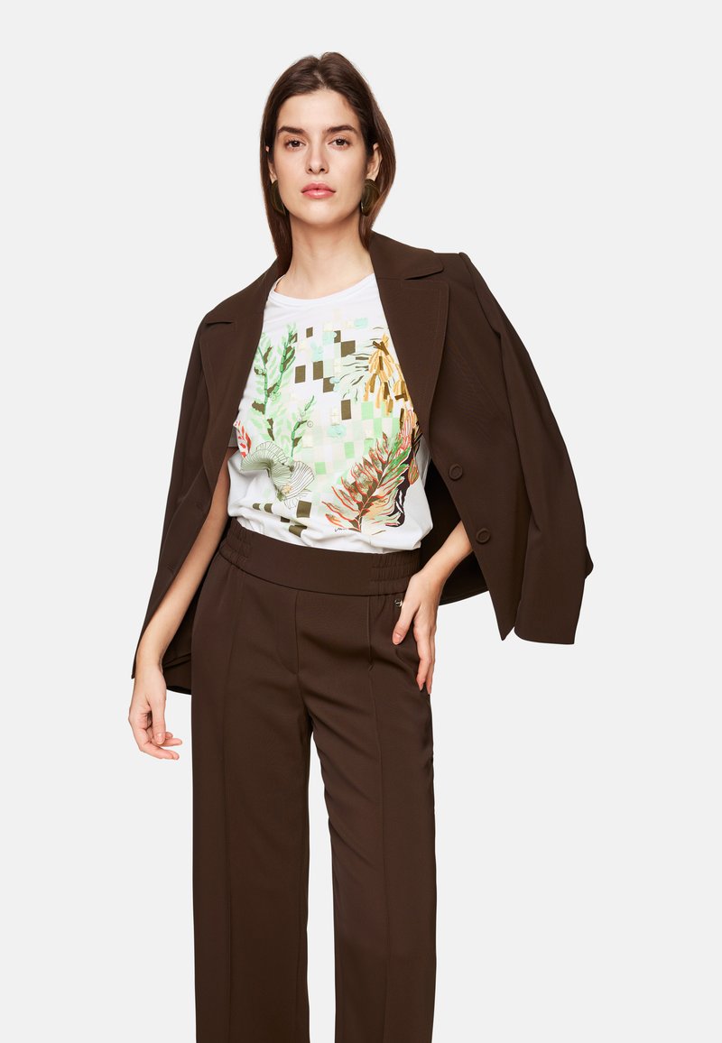 Brown tailored jacket over a white graphic t-shirt featuring botanical prints and geometric patterns, paired with brown wide-leg trousers.