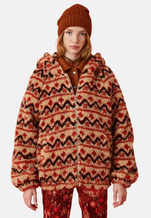 Flauschige, übergroße Jacke in Beige mit roten Zickzack- und Blumenmustern, ausgestattet mit einem Reißverschluss vorne und einer Kapuze. Kombiniert mit roten gemusterten Hosen.