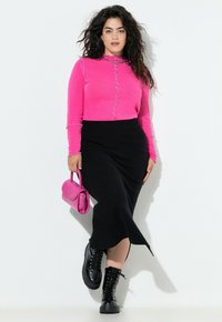 Vrouw met donker golvend haar, gekleed in een felroze lange mouw top, zwarte midi rok, zwarte veterschoenen, en houdt een roze handtas vast, terwijl ze tegen een witte muur staat.