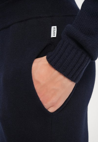 Marineblå strikkegenser med ribbestrikket mansjett, sidelomme og en liten merkeetikett. Stoffet har en myk, strukturert overflate.