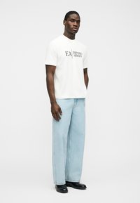 Man staat en draagt een wit Emporio Armani T-shirt, lichtblauwe wijde jeans en zwarte schoenen tegen een effen witte achtergrond.