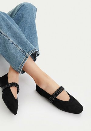 Füße in schwarzen Mary Jane Ballerinas aus Wildleder mit gerafften Riemen, kombiniert mit hellblauen Jeans vor weißem Hintergrund.