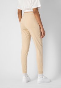 Beige Jogginghosen aus weichem Stoff mit einer Gesäßtasche und elastischem Bund. Dazu weiße Turnschuhe und gerippte Bündchen.