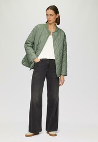 Gequilted groene jack met een ronde kraag, gedragen over een witte top, gecombineerd met donkerblauwe wijde jeans en beige schoenen met bruine zolen.