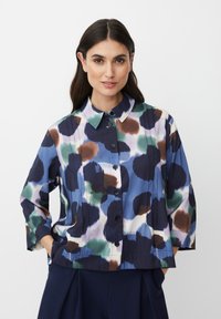 Donna con capelli scuri che indossa una camicetta a bottoni a fantasia multicolore e pantaloni scuri, mani in tasca, su uno sfondo chiaro e semplice.