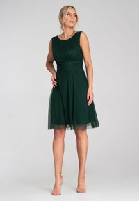 Grünes ärmelloses Kleid mit tailliertem Oberteil und schwingendem Rock, aus leichtem, fließendem Material. Hat eine glatte Textur und eine geraffte Vorderseite.