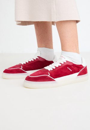 Person trägt weiße Socken und rote Wildledersneaker mit weißen Sohlen und Schnürsenkeln, steht auf einem hellen Boden mit beigefarbenen Hosen, die oberhalb der Knöchel gekürzt sind.