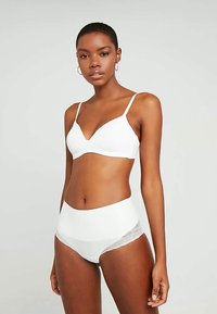 Mulher a usar um soutien branco e cuecas de cintura alta com rendas, de pé contra um fundo cinza claro e simples.