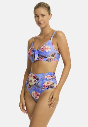 Modelka pozuje w żywej, niebieskiej bikini w kwiaty z wiązaniem z przodu. Jasne kwiaty i wysoki stan dolnej części podkreślają styl plażowy.