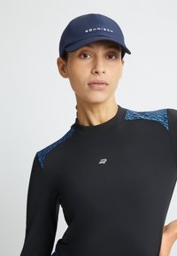 Cappellino da baseball navy con logo, abbinato a una maglietta nera a maniche lunghe con dettagli a motivi blu sulle spalle, dalla texture liscia.