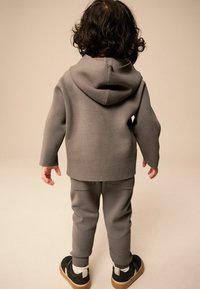 Grå huva-tröja och matchande byxor i textil, kombinerat med svarta skor och vita strumpor. Sedd från baksidan.