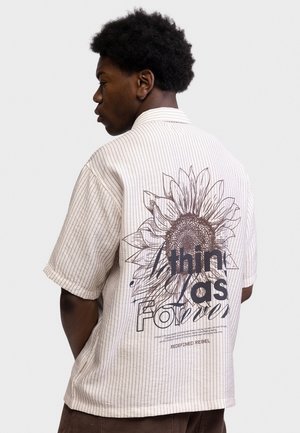 Person mit kurzen lockigen Haaren, die ein beiges Kurzarmhemd mit dünnen Streifen und einem großen Sonnenblumen- und Textdesign auf dem Rücken trägt.