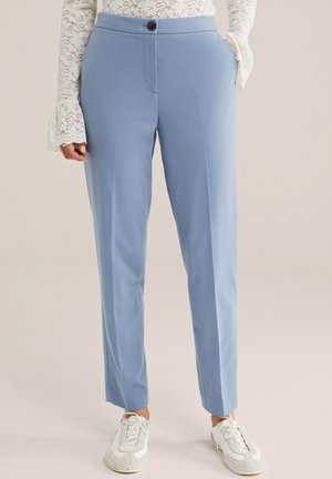 Femme portant un pantalon bleu clair ajusté avec bouton et poche à l'avant, associé à un haut en dentelle blanche à manches longues et des baskets blanches.