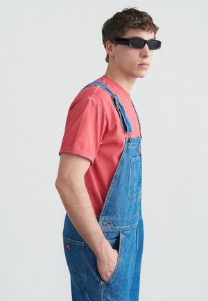 Jonge man draagt zwarte rechthoekige zonnebril, rood T-shirt en blauwe spijkerbroek met bretels, staat zijwaarts met hand in zak tegen witte achtergrond.