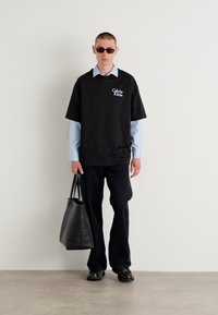 T-shirt noire oversize avec logo Calvin Klein, superposée à une chemise bleu clair, associée à un pantalon foncé et un tote bag noir.