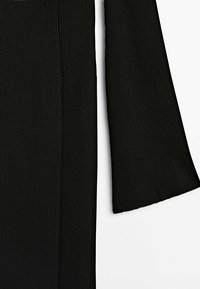 Gros plan d'un vêtement noir en maille côtelée à manches longues montrant la texture du tissu et le détail de la couture de la manche sur fond blanc.