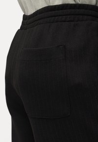 Gros plan sur un pantalon en tissu noir texturé avec une taille élastique et une poche arrière appliquée, porté avec un haut gris clair rentré dedans.