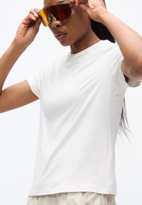 Γυναίκα που φοράει απλό λευκό μπλουζάκι προσαρμόζοντας μεγάλα αθλητικά γυαλιά ηλίου με πορτοκαλί απόχρωση σε λευκό φόντο.