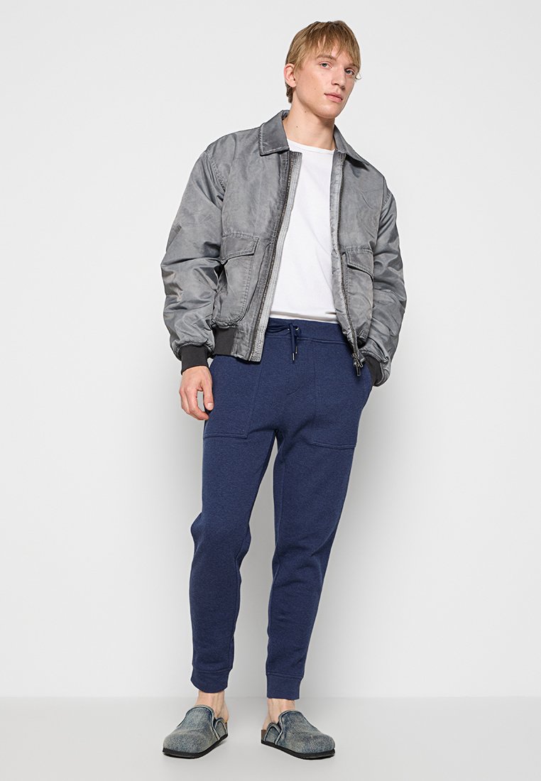 Szara kurtka bomberka z zamkiem z przodu i dwiema kieszeniami, w połączeniu z ciemnoniebieskimi spodniami dresowymi i szarymi butami wsuwanymi. Prosty, codzienny styl.