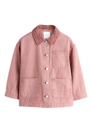 Ung pige i lyserød jakke og vide jeans læner sig op ad en brun murstensbygning med store vinduer på en byfortov.