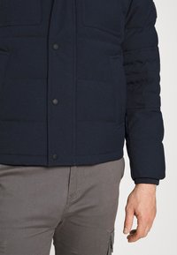 Granatowa pikowana kurtka z guzikami z przodu, dwoma kieszeniami na piersi i ściągaczami na mankietach. Jednokolorowy materiał o gładkiej fakturze. Szare spodnie.