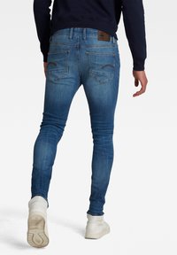 Personne marchant de dos portant un jean bleu slim, des baskets blanches et un haut sombre à manches longues avec des poches arrière visibles et des détails de couture.