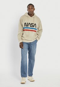 Beżowa bluza z kapturem z czarnym logo "NASA" oraz paskami w kolorze niebieskim i czerwonym. Połączona z jasnoniebieskimi dżinsami i beżowymi trampkami.