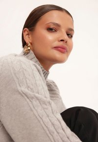 Szary sweter z wysokim kołnierzem i długimi rękawami, wykonany z dzianiny w warkocze, w połączeniu z czarnymi spodniami z materiału. Złote kolczyki w kształcie kół podkreślają stylizację.