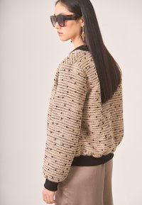 Suéter de textura beige con rayas negras y acentos metálicos, que presenta un cuello y puños acanalados. El modelo lleva gafas de sol oscuras.