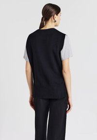 Top senza maniche nera in maglia con una finitura testurizzata, dotata di scollatura rotonda e orlo posteriore leggermente più lungo. Abbinato a una camicia grigia a maniche corte.