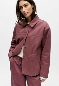 Kobieta ubrana w fioletowawy zapinany na guziki żakiet z kołnierzem i pasujące spodnie, ręce w kieszeniach, pod spodem kremowy sweter dzianinowy.