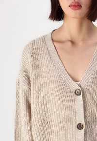 Beige stickad cardigan med V-ringning, långa ärmar och två bruna knappar fram. Texturerat mönster ger djup åt tyget.